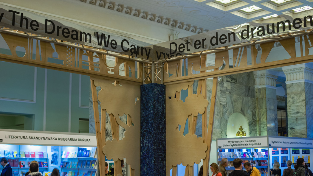 Part of a wooden stand at a book fair. At the top it says "The dream we carry" and "Det er den draumen". Small stands of polish publishers in the background. 
